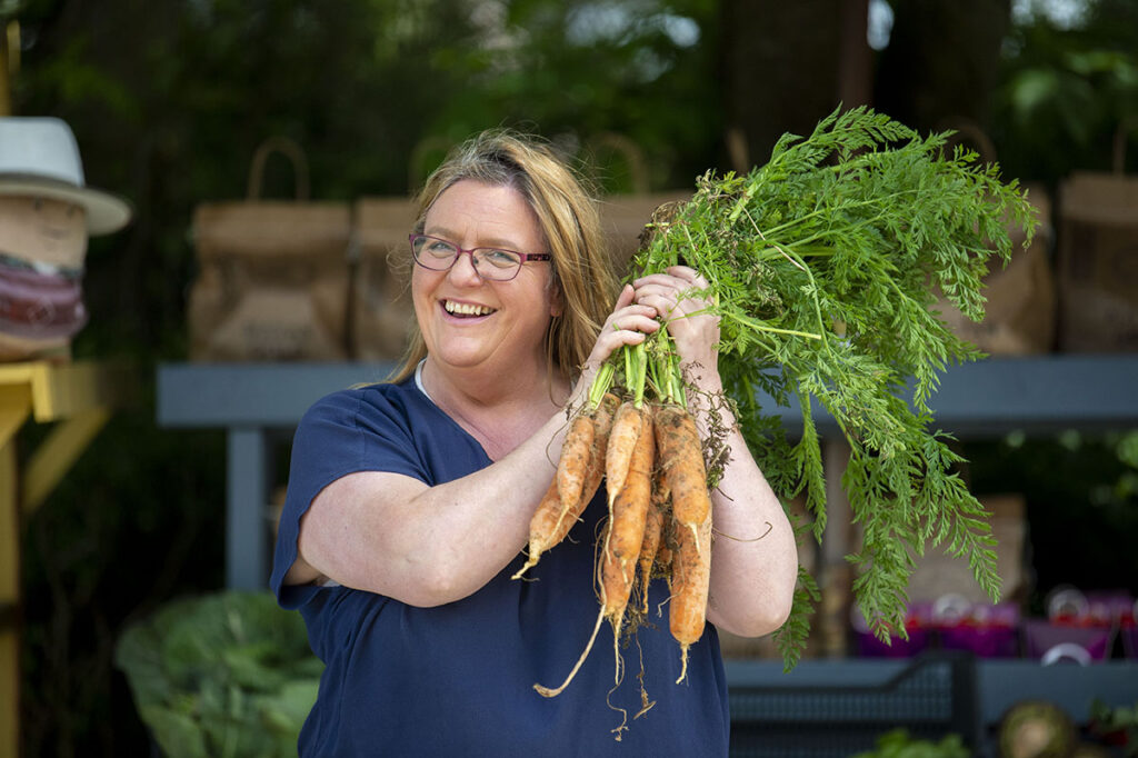 Maria Flynn of Ballymakenny Farm and the Spud Shack is holding a bunch of carrots grown locally. She is standing outside her Spud Shack and the sun is shining.  The photograph was taken by Solstice Media Ltd.